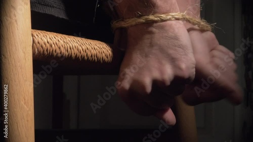 Man struggles with hands tied behind back to wooden chair inside a dark room with spotlight close up photo