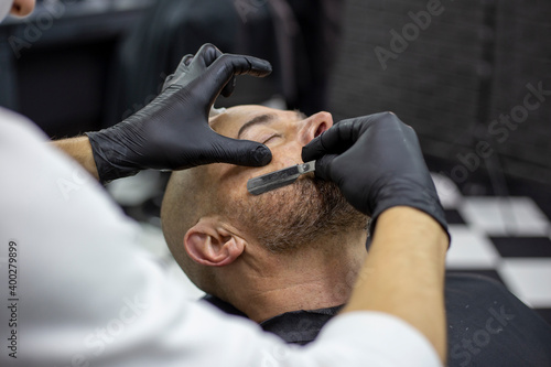 Hands of a hairdresser in black gloves shave the beard of a client with a straight razor in the salon