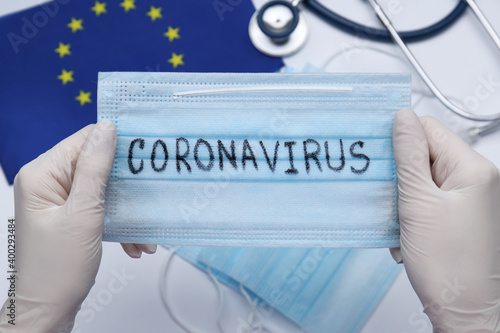 Doctor holding mask with word Coronavirus above medical items and European Union flag, closeup