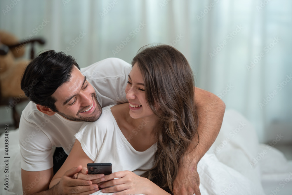Happy young relaxed couple using mobile phone and smiling while spending time in bed at home