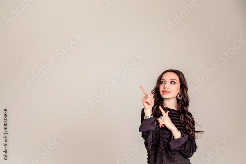 beautiful slender young girl with black long hair on an isolated beige and gray background. woman in brown trousers and black blouse shows with forefinger hand