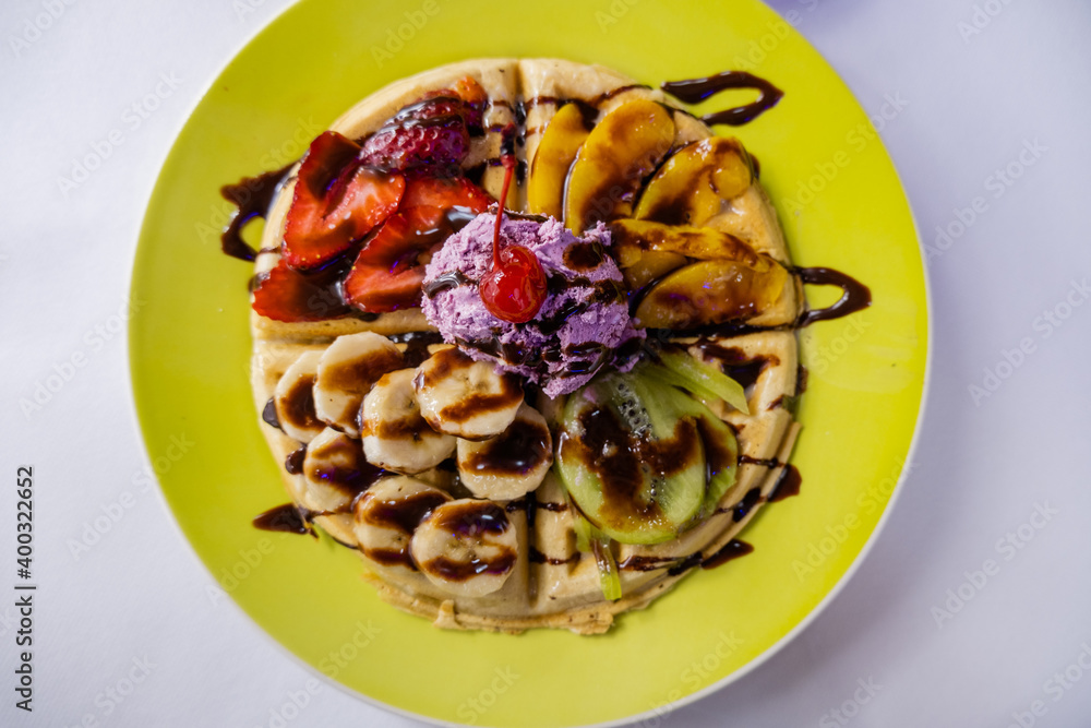 Waffle on a yellow plate with fruit slices and chocolate syrup on top