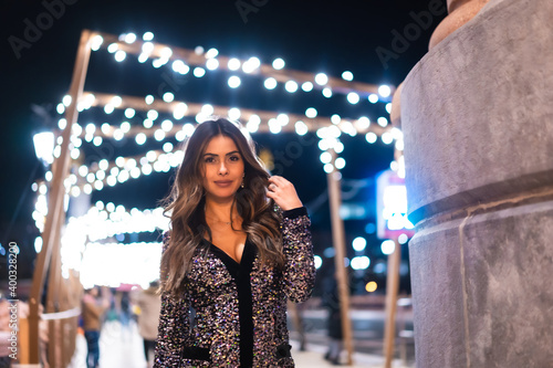 Christmas lifestyle. Portrait a young brunette Caucasian woman in a fashionable dress with sequins, smiling in the background city Christmas lights