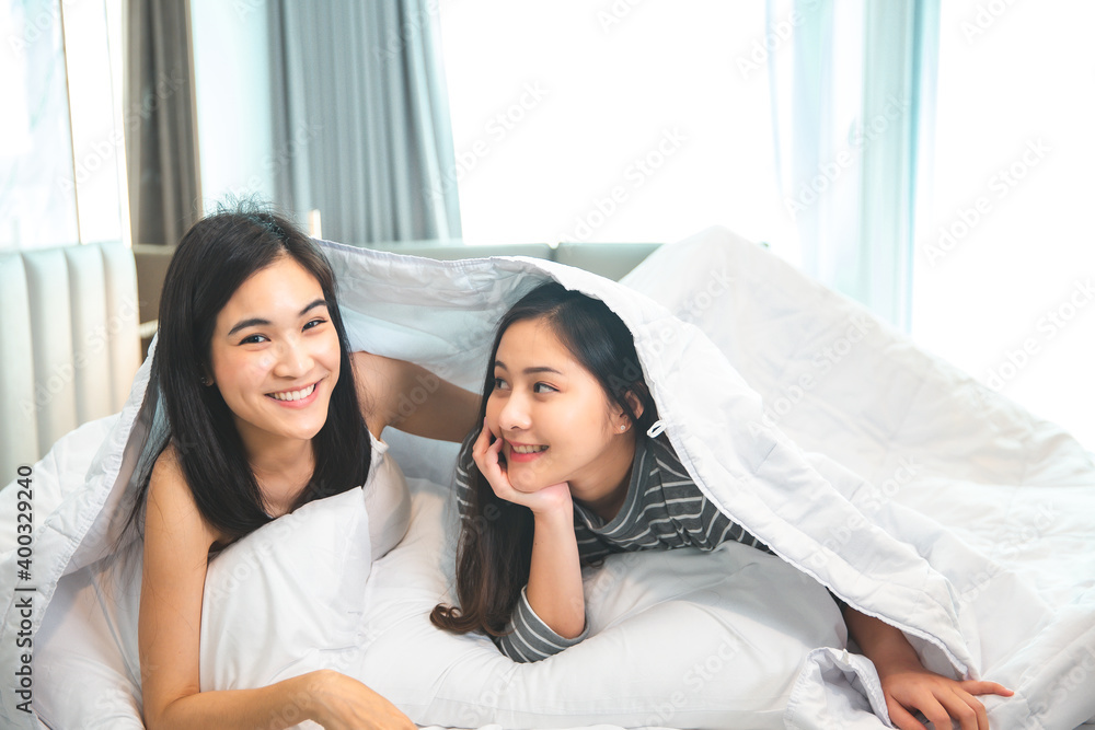 Two young asian cute women in bedroom at home lying on bed covered with blanket looking camera smiling cheerful. Two young asian cute women in bedroom at home lying on bed covered with blanket looking camera smiling cheerful.