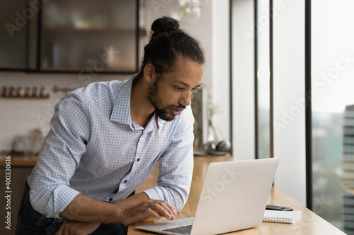 Young African American man look at laptop screen working online in office. Concentrated millennial biracial male employee busy using computer at workplace, browse surf wireless internet on device.