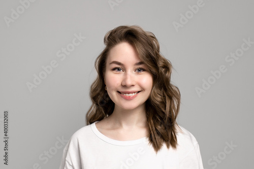 Portrait of happy smiling young woman looking at camera with natural everyday makeup, gray background, copy space. Beautiful caucasian girl with brown curly hairstyle. Cheerful expression face