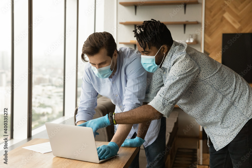 Multiracial male employees in facial masks and rubber gloves cooperate work on laptop in office. Diverse men in medical protective gear use computer during covid-19 pandemics. Corona virus concept. Multiracial male employees in facial masks and rubber gloves cooperate work on laptop in office. Diverse men in medical protective gear use computer during covid-19 pandemics. Corona virus concept.