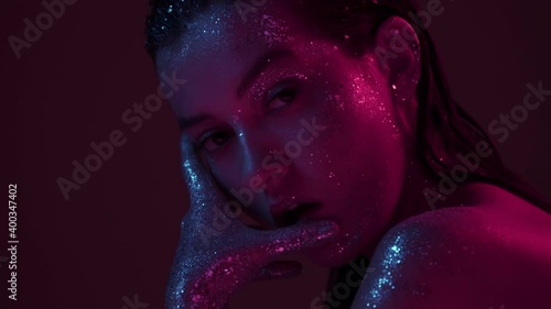 Sensual girl in sparkles in neon light posing in studio, sexy female with glittering make up and bare shoulder