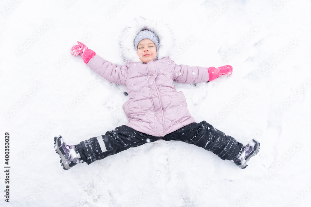 a cute little girl 4 years old in a pink down jacket and a hood lies in a snowdrift rejoicing in the snow. winter fun a cute little girl 4 years old in a pink down jacket and a hood lies in a snowdrift rejoicing in the snow. winter fun