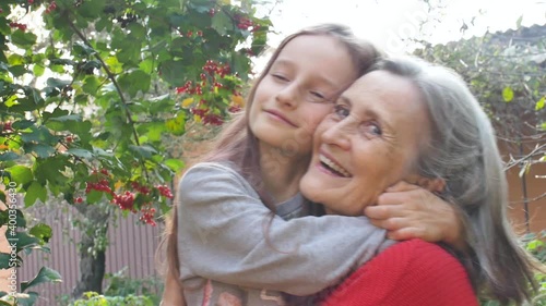 Senior grandmother with gray hair wearing red sweater with her little granddaughter are hugging in the garden and during sunny day outdoors, mother's day