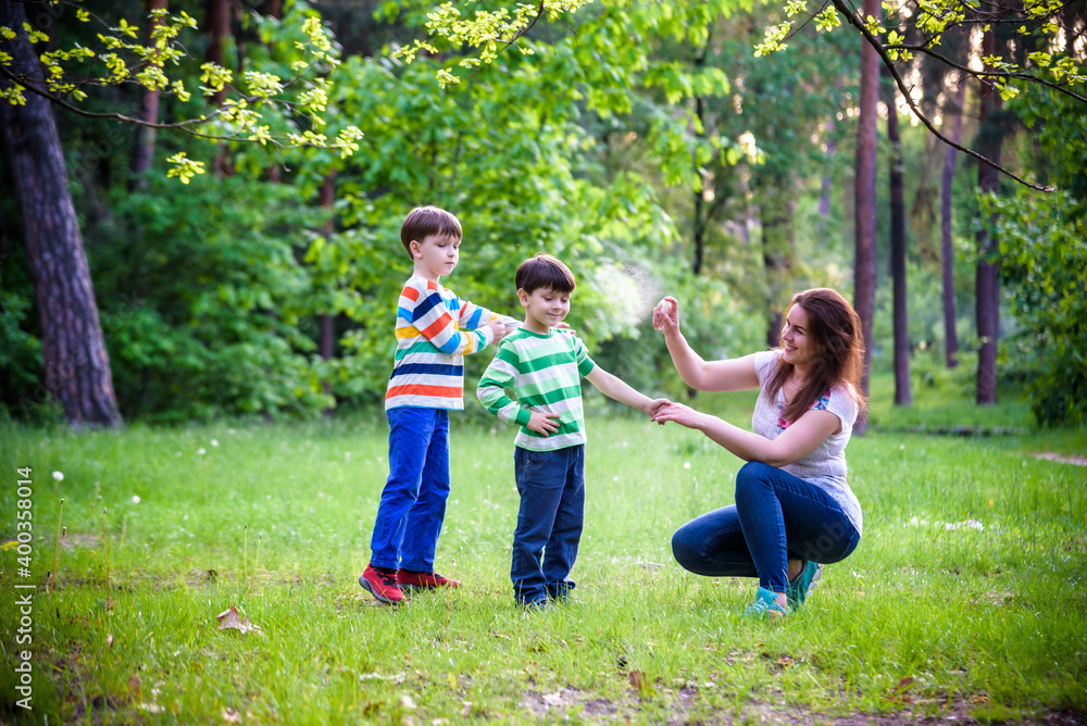 Fototapeta premium Young woman mother applying insect repellent to her two son before forest hike beautiful summer day or evening. Protecting children from biting insects at summer. Active leisure with kids