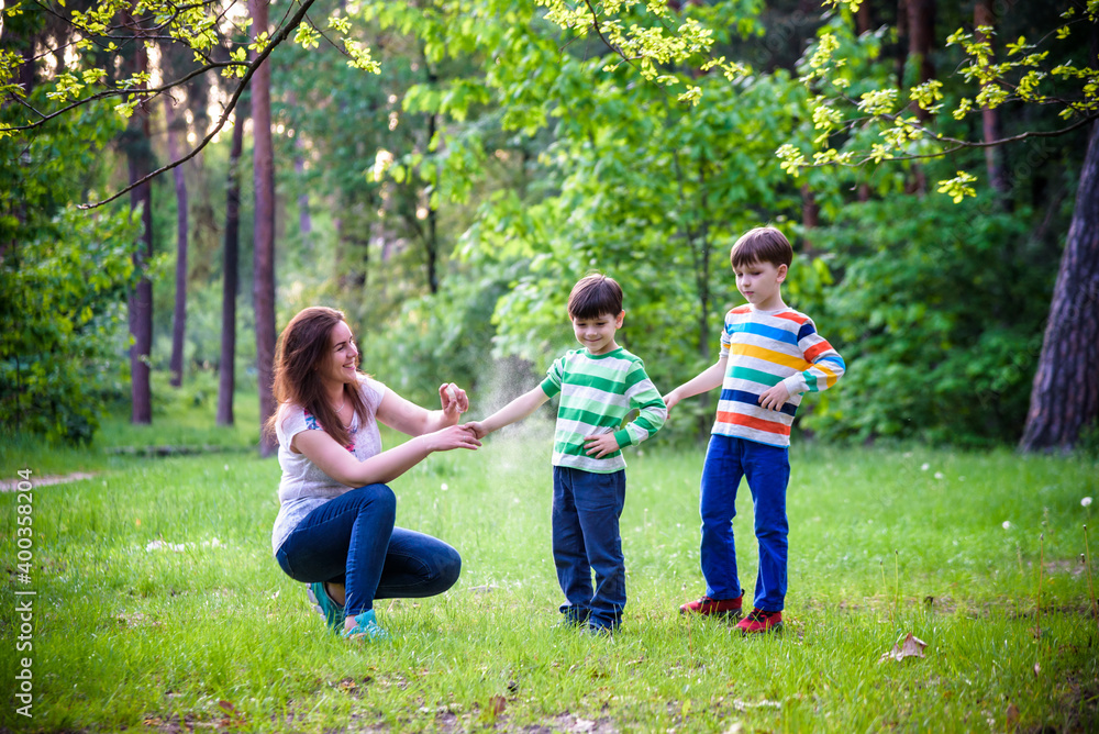 Fototapeta premium Young woman mother applying insect repellent to her two son before forest hike beautiful summer day or evening. Protecting children from biting insects at summer. Active leisure with kids