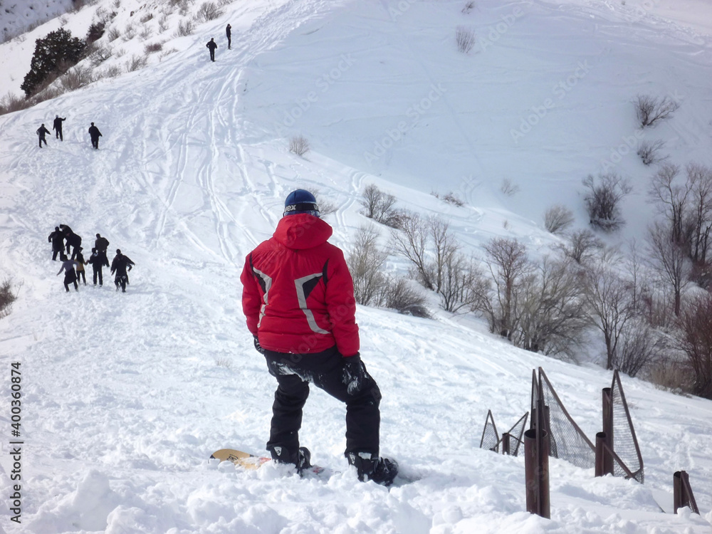 Guy in a red jacket stands on a snowboard on a mountain top in the snow, view from the back