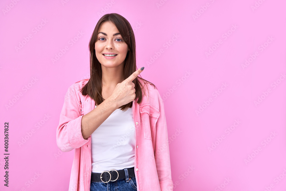 Pretty brunette girl in casual smiles and points finger aside at copy space, showing something amazing and looks at the camera isolated on pink background Pretty brunette girl in casual smiles and points finger aside at copy space, showing something amazing and looks at the camera isolated on pink background