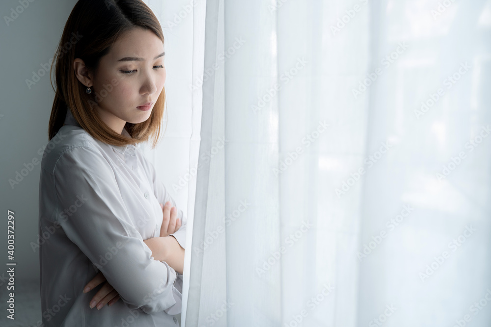 Depressed young Asian woman near window at home.