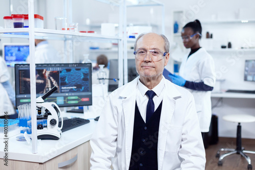 Senior male chemist looking at camera wearing protective glasses. Elderly scientist wearing lab coat working to develop a new medical vacine with african assistant in the background.