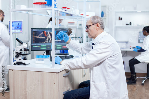 Senior scientist analysing and holding micobiology test tube in busy laboratory. Viorolog researcher in professional lab working to discover medical treatment.