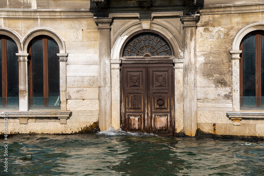 Venice, Italy. Close up of a waterfront bulding with wooden portal or door partlu submerged by the water. Water at very high level submerging steps and doorways and almost touching the windows.