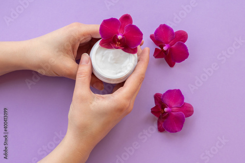 Young girls hands are holding soft white cream and magenta orchids.Moisturizing facial cream in a jar and blooming orchid flowers on purple background, skin care cosmetics.