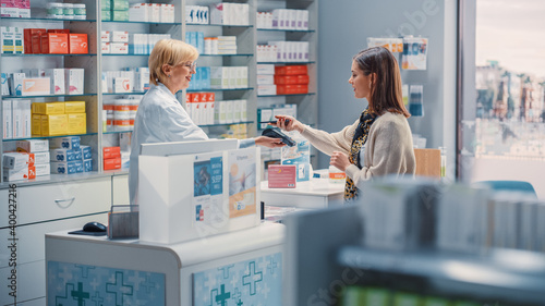 Pharmacy Drugstore Checkout Cashier Counter: Mature Female Pharmacist and Young Woman Using Contactless Payment NFC Smartphone to Buy Prescription Medicine, Vitamins, Beauty, Health Care Products photo