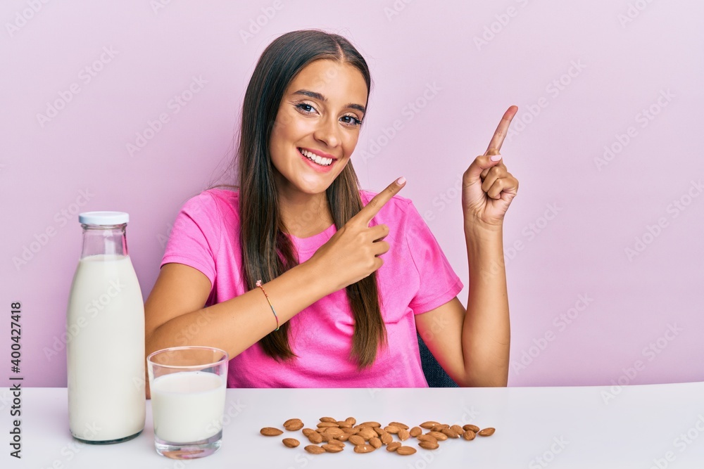 Young hispanic woman drinking healthy almond milk sitting on the table smiling and looking at the camera pointing with two hands and fingers to the side. Young hispanic woman drinking healthy almond milk sitting on the table smiling and looking at the camera pointing with two hands and fingers to the side.