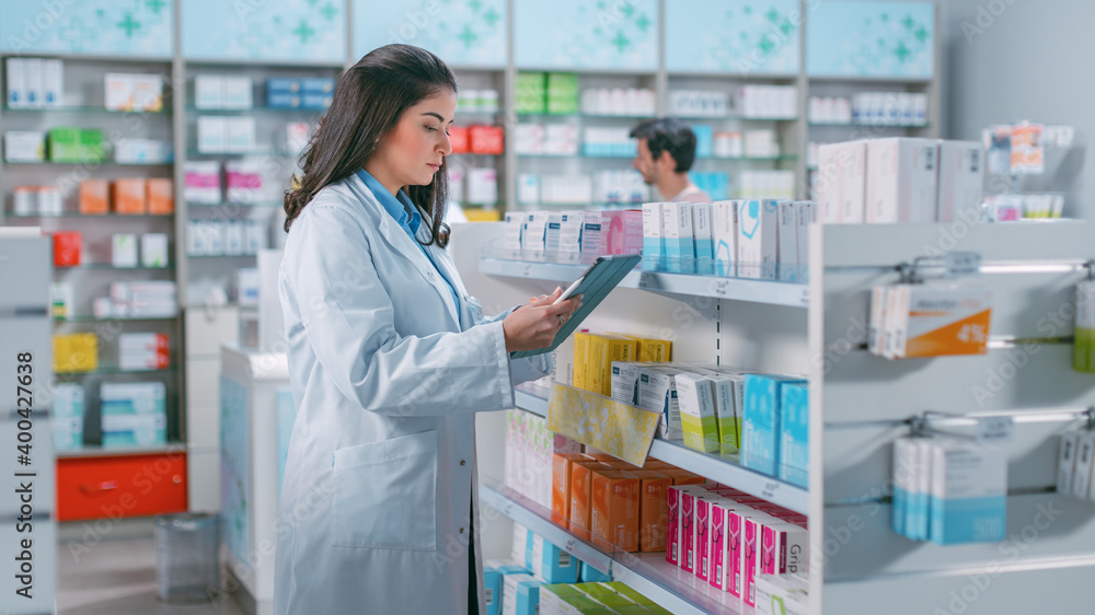 Pharmacy Drugstore: Beautiful Latina Pharmacist Uses Digital Tablet Computer, Checks Inventory of Medicine, Drugs, Vitamins, Health Care Products on a Shelf. Professional Pharmacist in Pharma Store