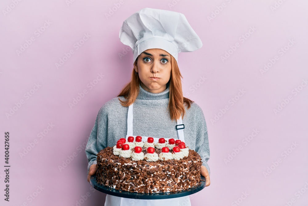 Hispanic young chef woman holding chocolate cake puffing cheeks with funny face. mouth inflated with air, catching air. Hispanic young chef woman holding chocolate cake puffing cheeks with funny face. mouth inflated with air, catching air.