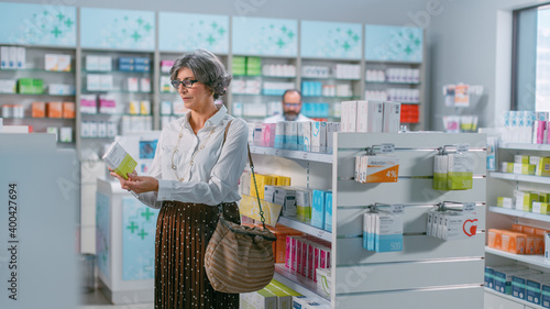 Pharmacy Drugstore: Portrait of a Beautiful Senior Woman Choosing to Buy Medicine, Drugs, Vitamins. Professional Pharmacist in Modern Pharma Store Shelves with Health Care Products.