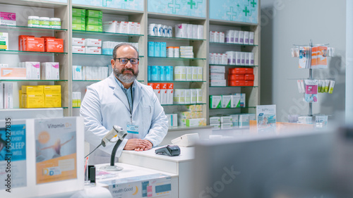 Pharmacy Drugstore Checkout Cashier Counter: Portrait of Experienced Mature Latin Pharmacist Looks at the Camera Smilingly. Pharma Store with Medicine, Drugs, Vitamins, Health Care Products.