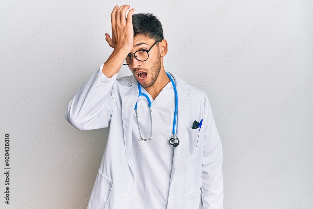 Young handsome man wearing doctor uniform and stethoscope surprised with hand on head for mistake, remember error. forgot, bad memory concept.