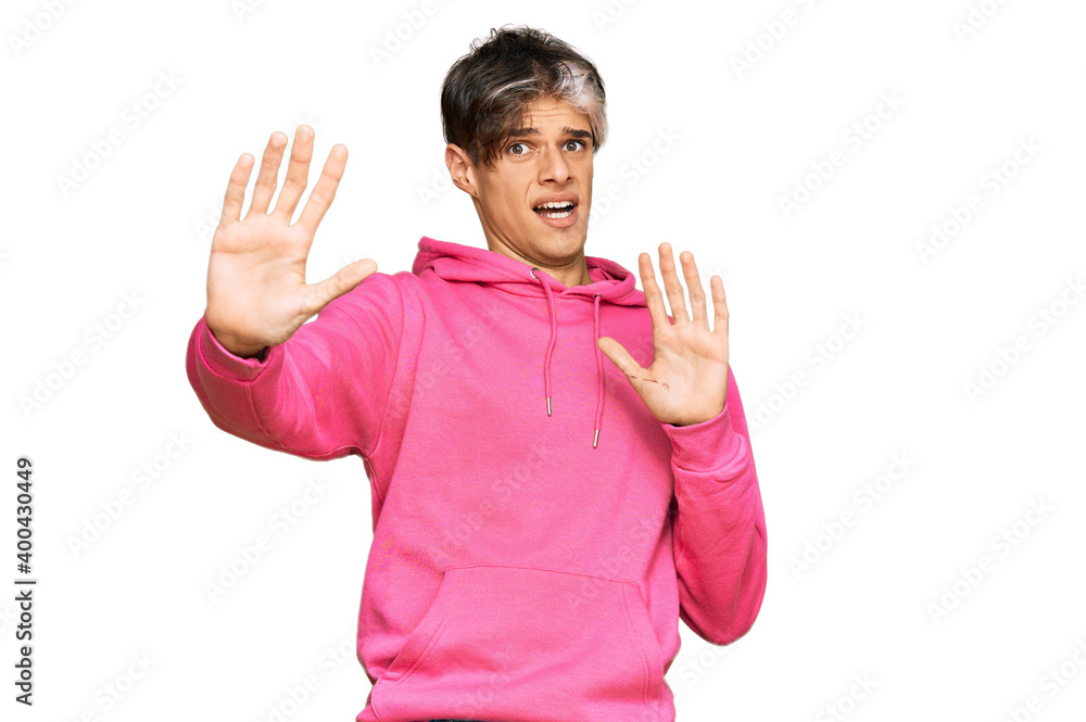 Young hispanic man wearing casual pink sweatshirt afraid and terrified with fear expression stop gesture with hands, shouting in shock. panic concept. Young hispanic man wearing casual pink sweatshirt afraid and terrified with fear expression stop gesture with hands, shouting in shock. panic concept.