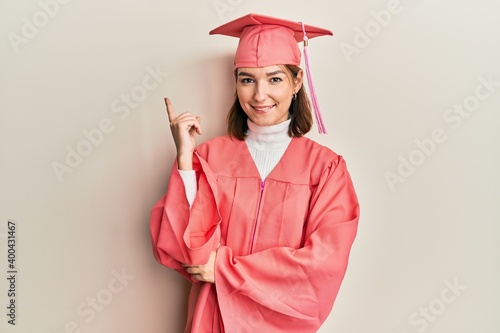 Young caucasian woman wearing graduation cap and ceremony robe with a big smile on face, pointing with hand and finger to the side looking at the camera.