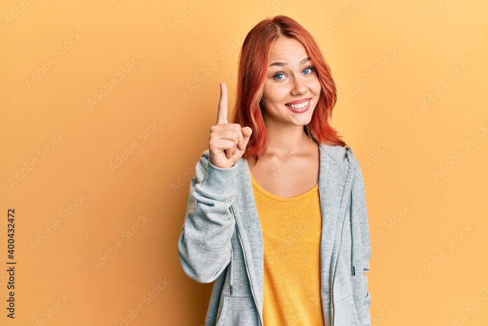 Young beautiful redhead woman wearing casual sporty sweatshirt pointing finger up with successful idea. exited and happy. number one. Young beautiful redhead woman wearing casual sporty sweatshirt pointing finger up with successful idea. exited and happy. number one.