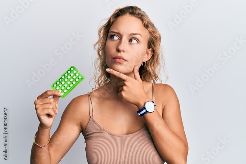 Beautiful caucasian woman holding birth control pills serious face thinking about question with hand on chin, thoughtful about confusing idea