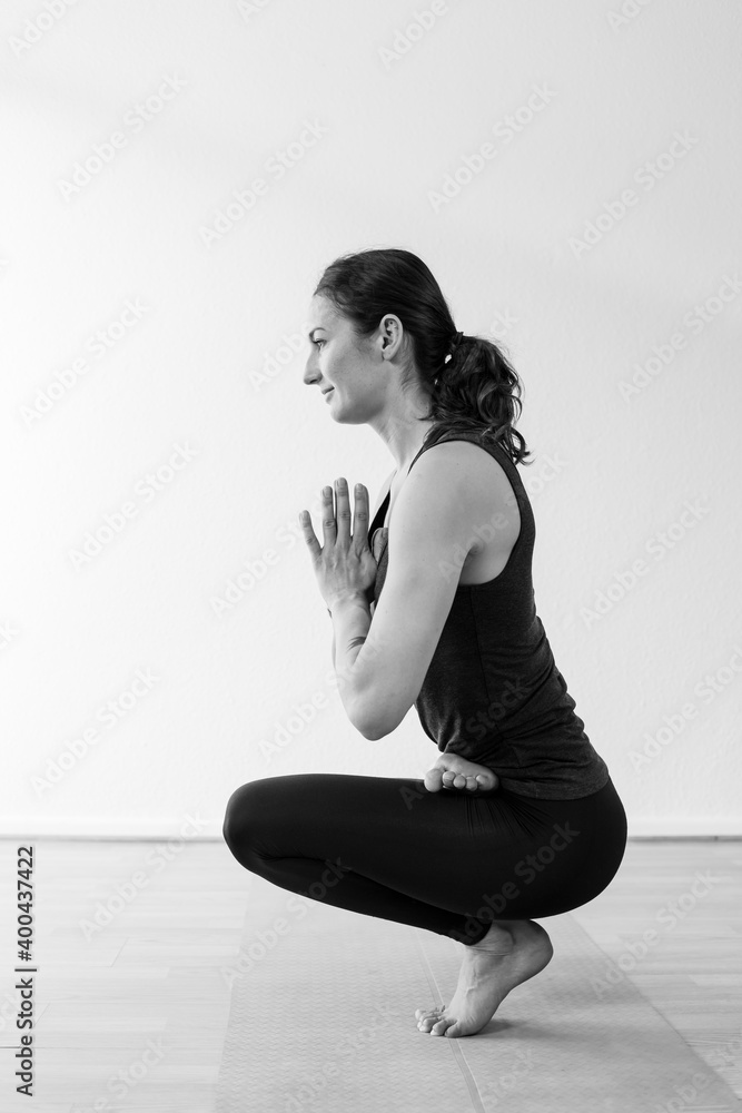 Fototapeta premium Side view of the tiptoe asana or yoga pose, with hands together at center of chest. A brunette woman in her 30’s practicing yoga at home.