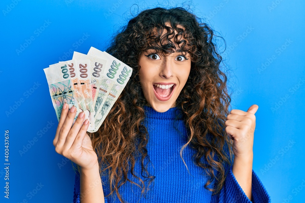 Young hispanic girl holding czech koruna banknotes pointing thumb up to the side smiling happy with open mouth Young hispanic girl holding czech koruna banknotes pointing thumb up to the side smiling happy with open mouth