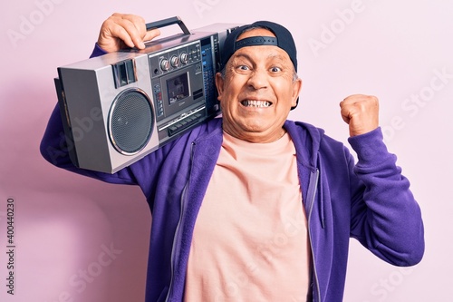Senior handsome grey-haired modern man listening to music using vintage boombox screaming proud, celebrating victory and success very excited with raised arm