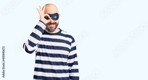 Young handsome man wearing burglar mask doing ok gesture with hand smiling, eye looking through fingers with happy face. © Krakenimages.com
