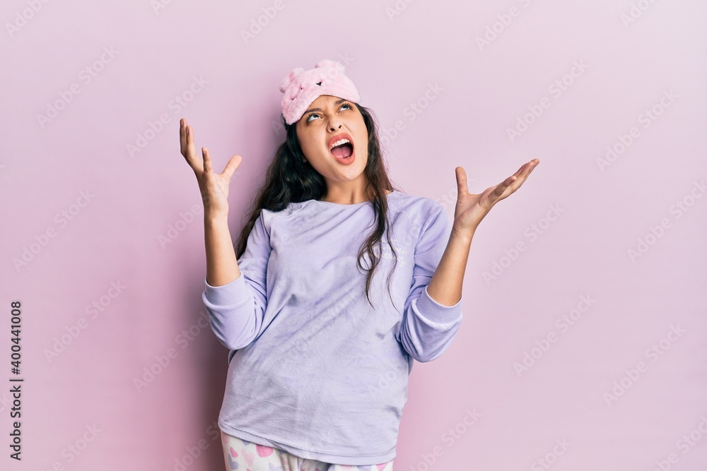 Beautiful middle eastern woman wearing sleep mask and pajama crazy and mad shouting and yelling with aggressive expression and arms raised. frustration concept. Beautiful middle eastern woman wearing sleep mask and pajama crazy and mad shouting and yelling with aggressive expression and arms raised. frustration concept.