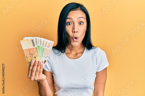 Beautiful hispanic woman holding south korean won banknotes scared and amazed with open mouth for surprise, disbelief face