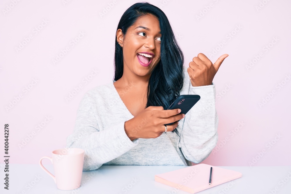 Beautiful latin young woman with long hair using smartphone sitting on the table pointing thumb up to the side smiling happy with open mouth Beautiful latin young woman with long hair using smartphone sitting on the table pointing thumb up to the side smiling happy with open mouth