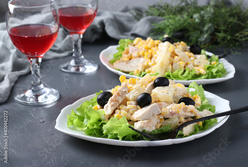 Festive salad with pineapple  baked chicken  corn and black olives on white plates and two glass of red wine on grey background. Horizontal format
