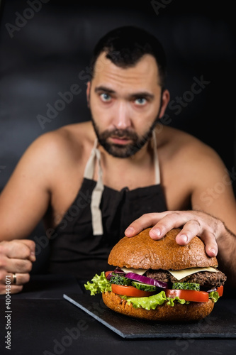 Cook in an apron on a naked body. Big and delicious Burger. Fast food.