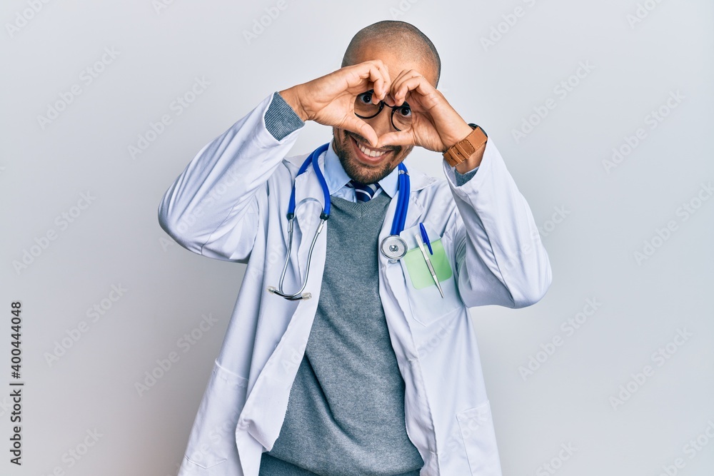 Hispanic adult man wearing doctor uniform and stethoscope doing heart shape with hand and fingers smiling looking through sign Hispanic adult man wearing doctor uniform and stethoscope doing heart shape with hand and fingers smiling looking through sign