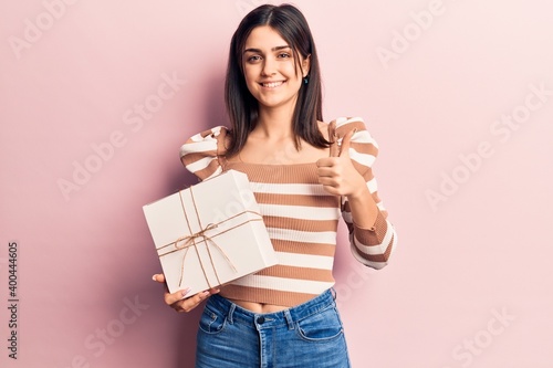 Young beautiful girl holding gift smiling happy and positive, thumb up doing excellent and approval sign