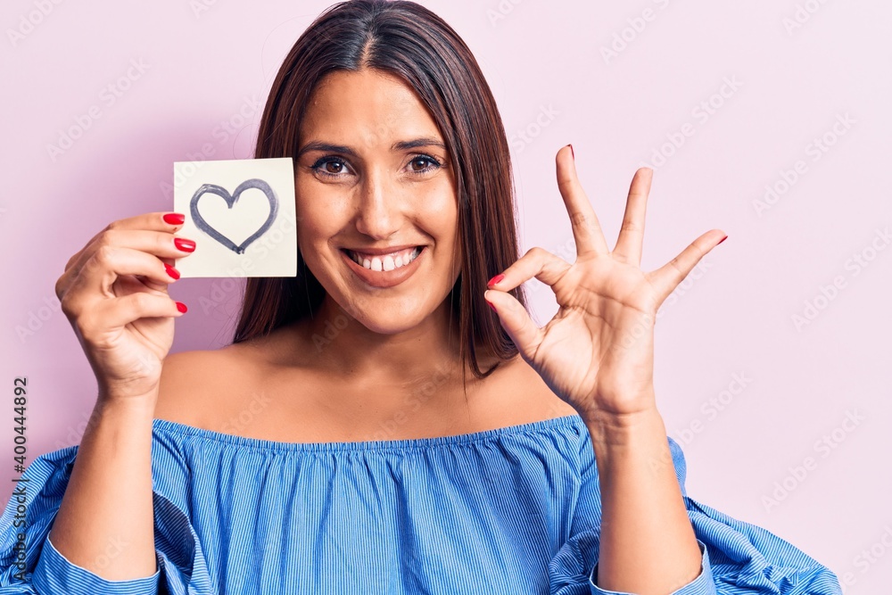 Young beautiful brunette woman holding reminder with heart doing ok sign with fingers, smiling friendly gesturing excellent symbol Young beautiful brunette woman holding reminder with heart doing ok sign with fingers, smiling friendly gesturing excellent symbol