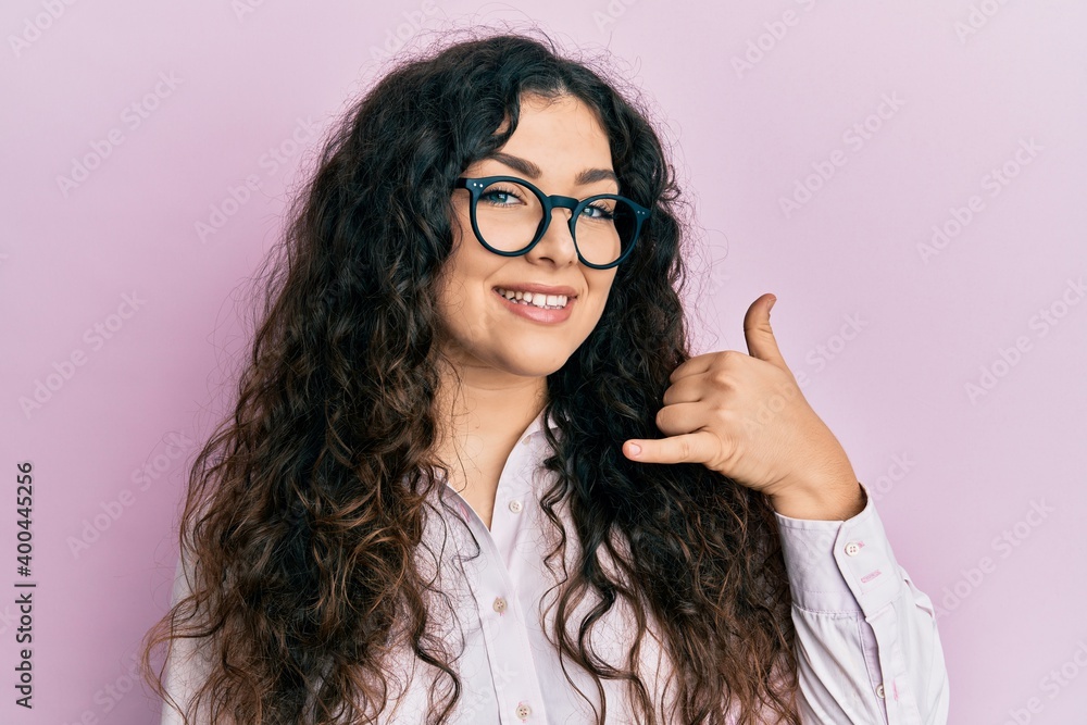 Obraz premium Young brunette woman with curly hair wearing casual clothes and glasses smiling doing phone gesture with hand and fingers like talking on the telephone. communicating concepts.