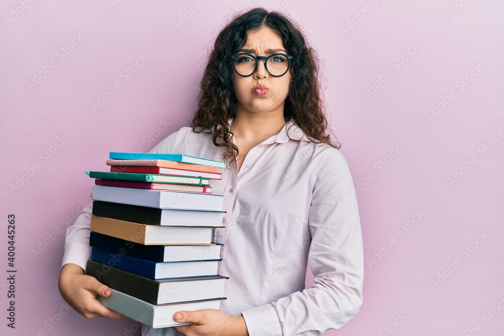 Young brunette woman with curly hair holding a pile of books puffing cheeks with funny face. mouth inflated with air, catching air. Young brunette woman with curly hair holding a pile of books puffing cheeks with funny face. mouth inflated with air, catching air.