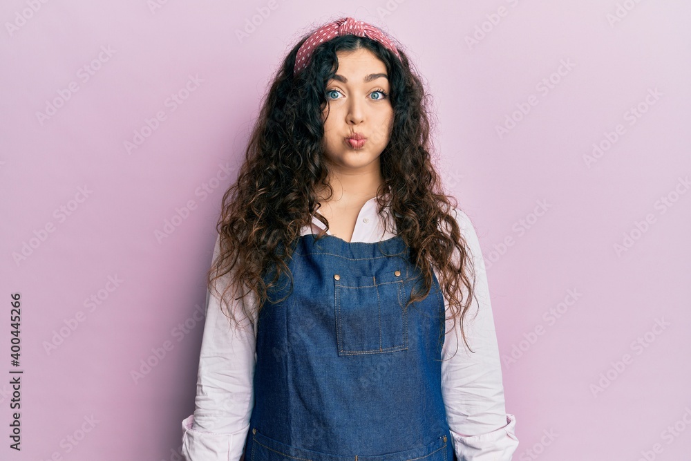 Young brunette woman with curly hair wearing cleaner apron and gloves puffing cheeks with funny face. mouth inflated with air, crazy expression. Young brunette woman with curly hair wearing cleaner apron and gloves puffing cheeks with funny face. mouth inflated with air, crazy expression.