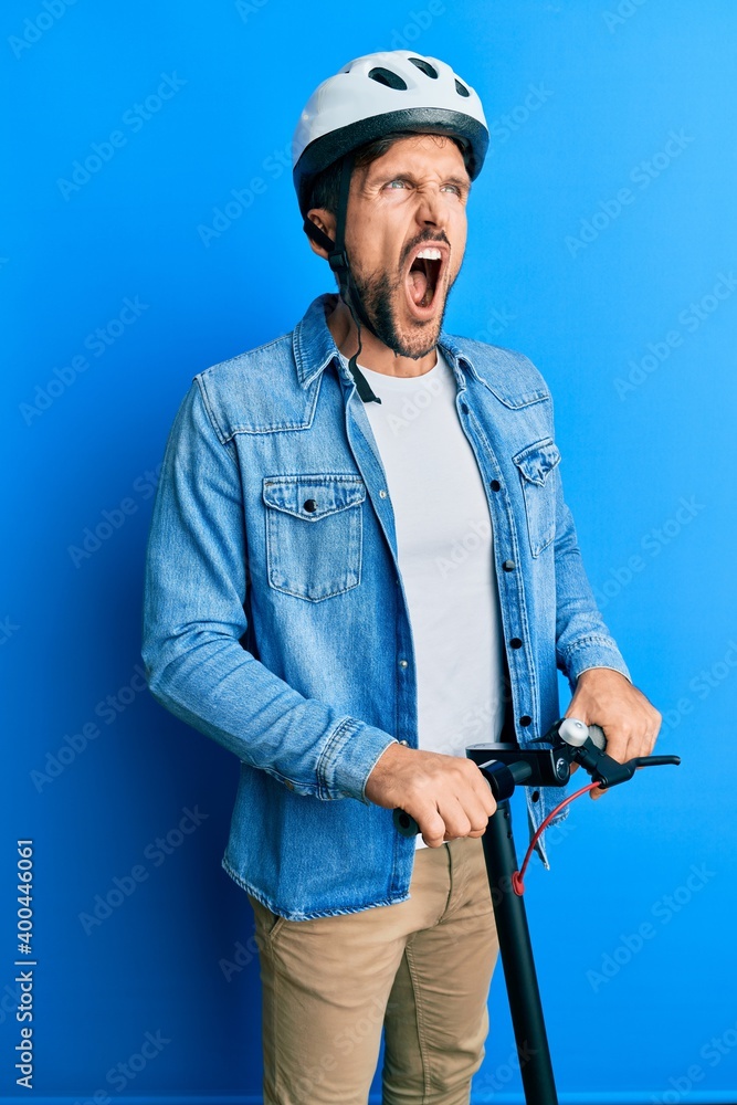 Young handsome man standing on modern scooter wearing helmet angry and mad screaming frustrated and furious, shouting with anger. rage and aggressive concept. Young handsome man standing on modern scooter wearing helmet angry and mad screaming frustrated and furious, shouting with anger. rage and aggressive concept.